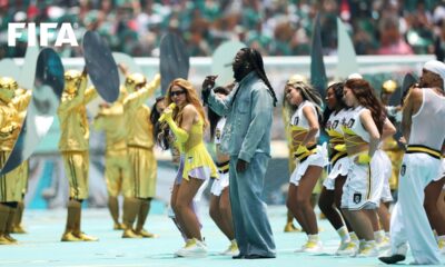 Burna Boy in a denim outfit and Shakira in a yellow dress singing together on stage with backup dancers at Estadio Azteca.