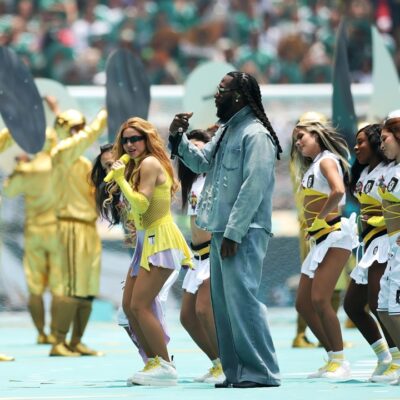 Burna Boy in a denim outfit and Shakira in a yellow dress singing together on stage with backup dancers at Estadio Azteca.