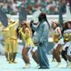 Burna Boy in a denim outfit and Shakira in a yellow dress singing together on stage with backup dancers at Estadio Azteca.