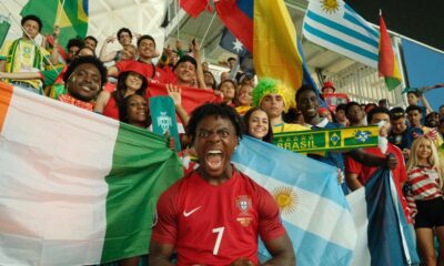 IShowSpeed wearing a red Portugal jersey with number 7, shouting excitedly in a stadium surrounded by diverse football fans holding flags from Nigeria, Argentina, Brazil, and Uruguay.