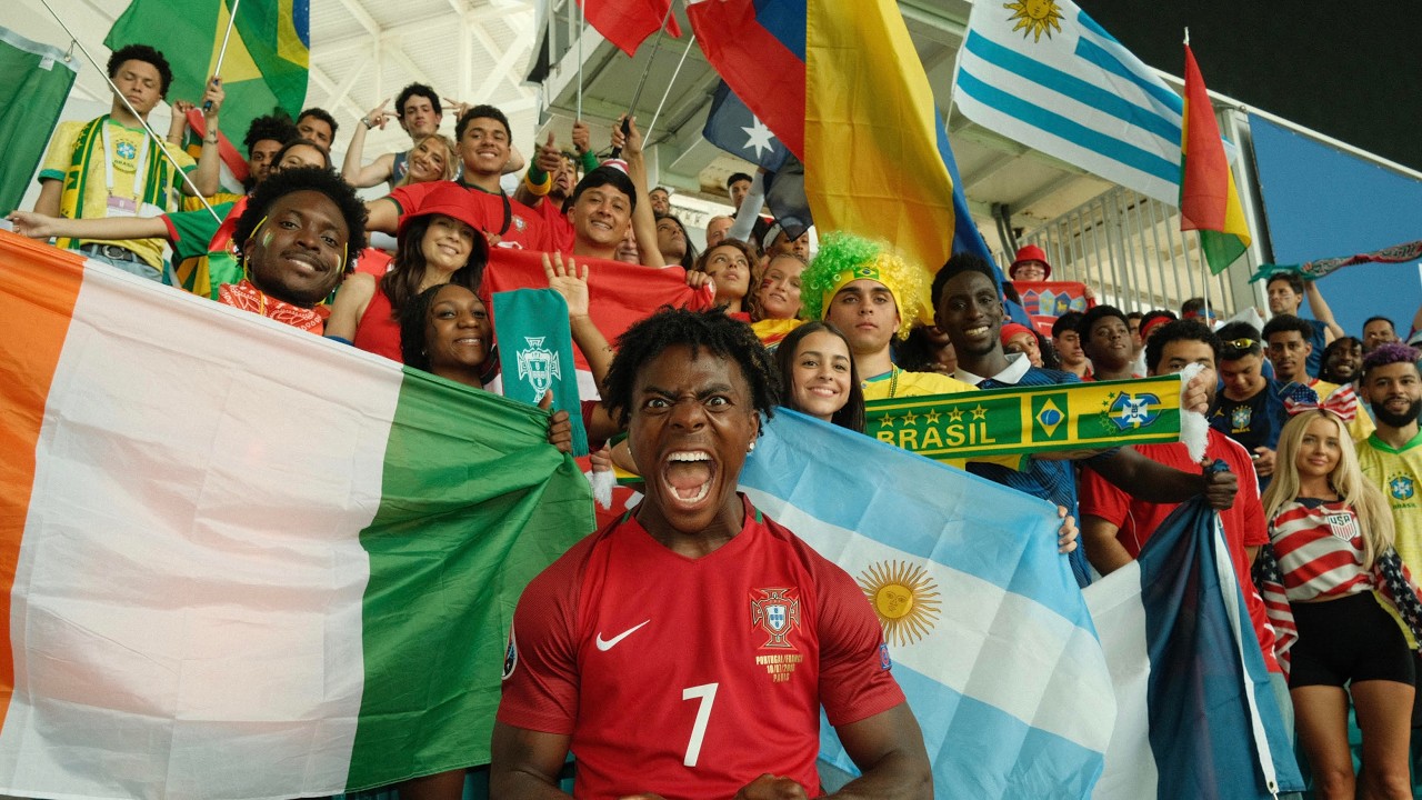 IShowSpeed wearing a red Portugal jersey with number 7, shouting excitedly in a stadium surrounded by diverse football fans holding flags from Nigeria, Argentina, Brazil, and Uruguay.