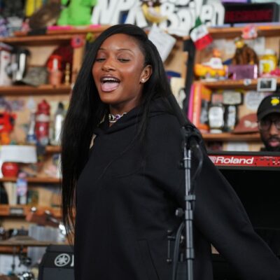 Nigerian singer Ayra Starr singing into a microphone during her NPR Tiny Desk Concert performance in a black sweatsuit with her live band in the background.