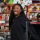 Nigerian singer Ayra Starr singing into a microphone during her NPR Tiny Desk Concert performance in a black sweatsuit with her live band in the background.