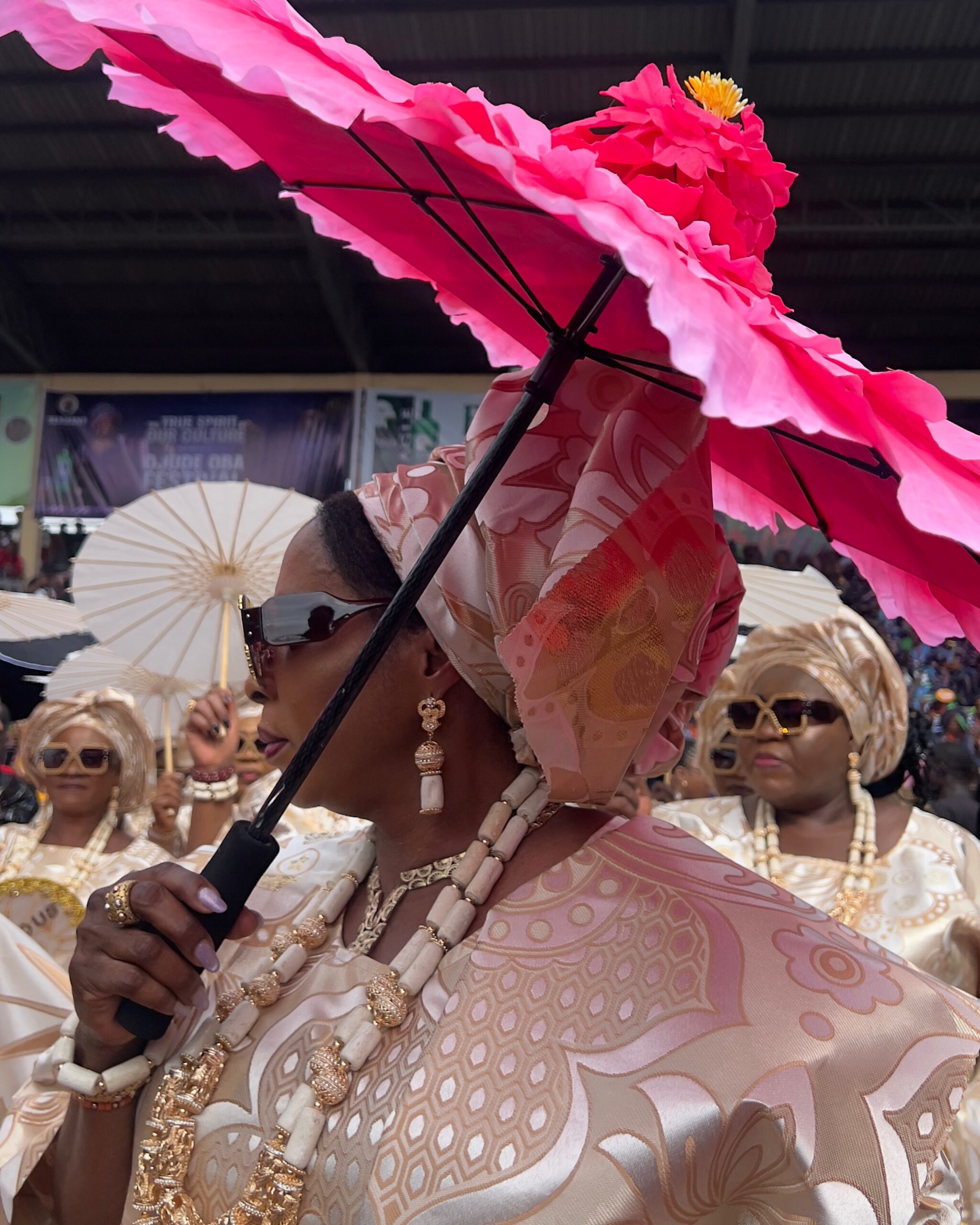 Profile view of a woman holding a vibrant pink flower-shaped parasol umbrella, wearing a patterned cream and pink damask gele, sunglasses, and layered traditional beads at the Ojude Oba Festival