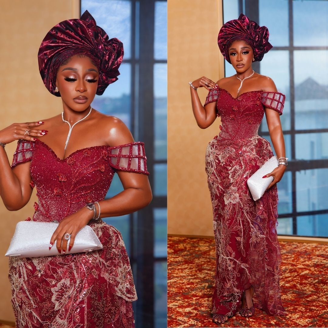 Ini Edo wearing an off-the-shoulder crimson net lace gown embroidered with gold floral threadwork, crystal beads, lattice-pattern sleeves, and a multi-pleated burgundy gele.