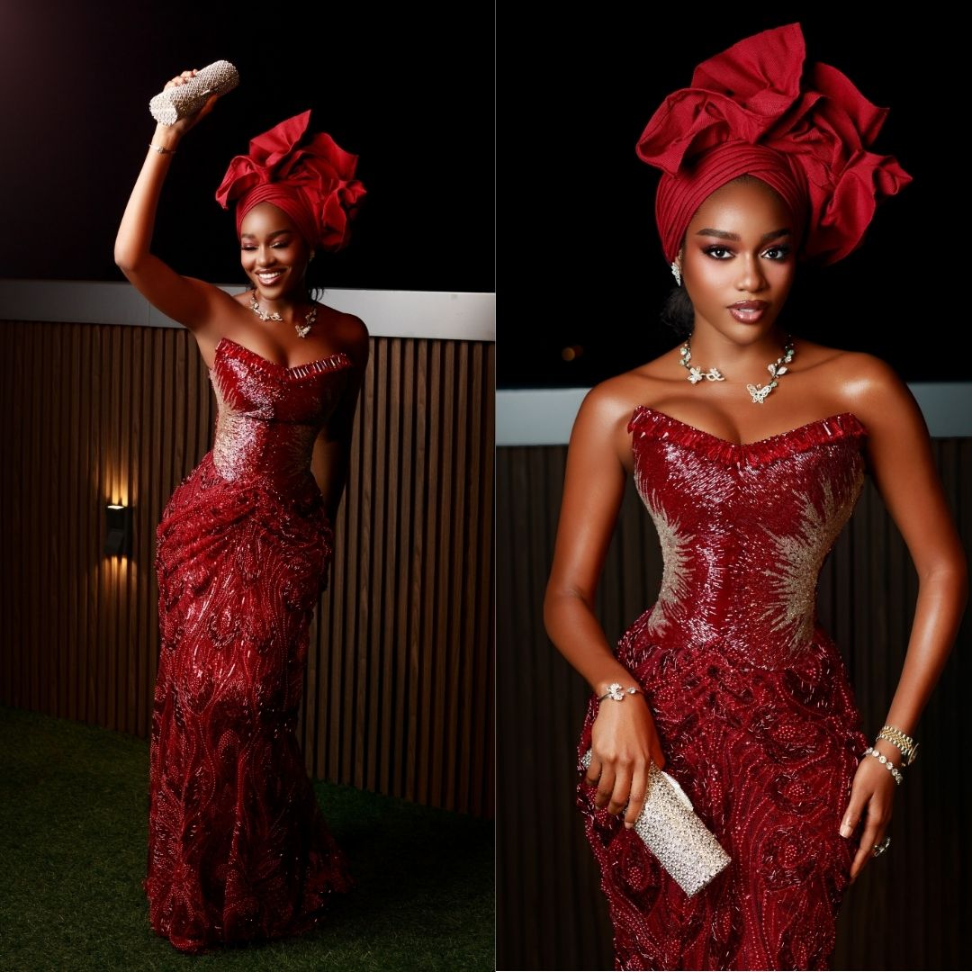 Beauty Tukura wearing a strapless crimson corset gown with silver metallic bust detailing, draped lace skirt, dramatic rose-shaped gele, and crystal clutch bag