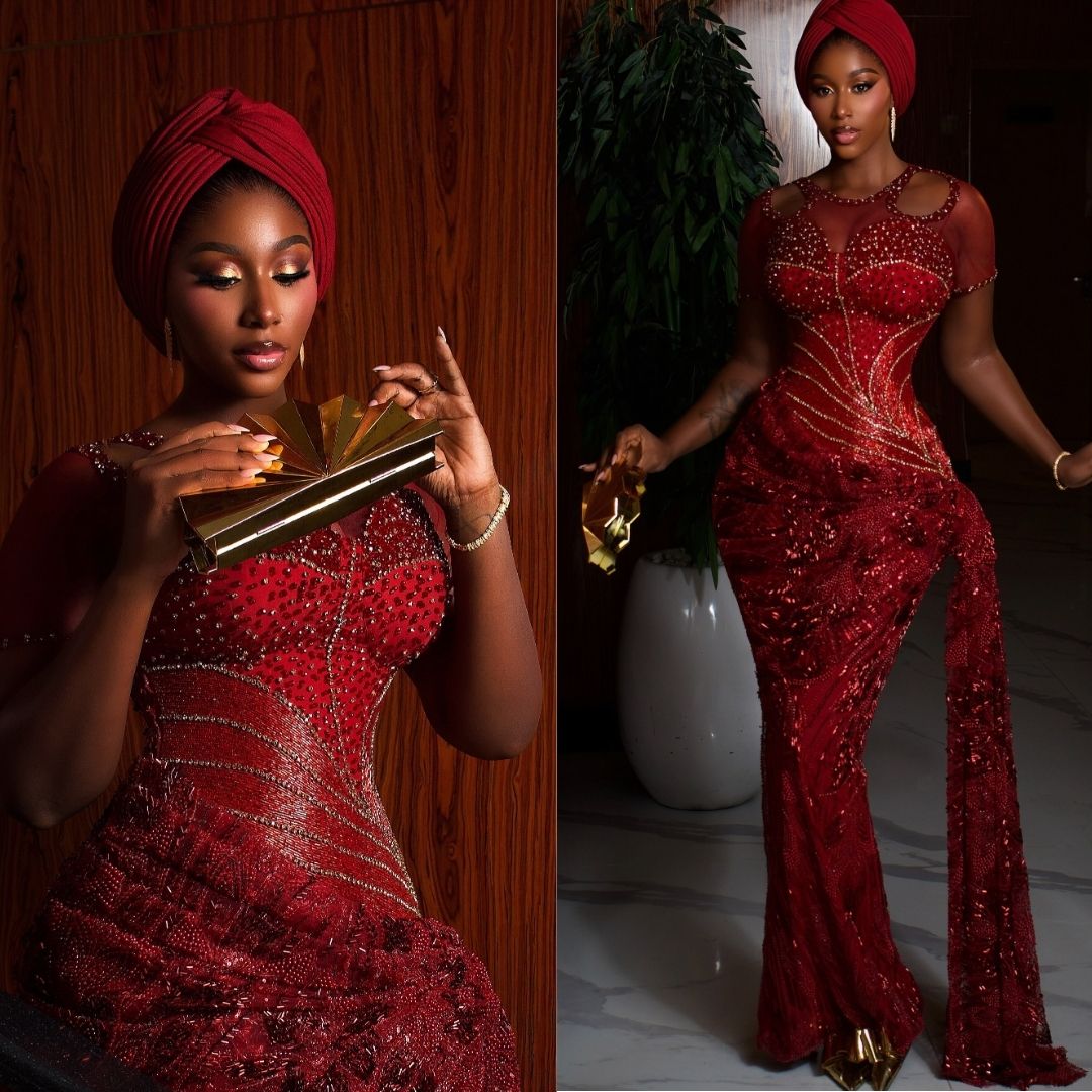 Nelly Mbonu in a short-sleeved deep burgundy corset gown with geometric crystal lines, sheer chest panel, red turban headwrap, and gold fan clutch bag.