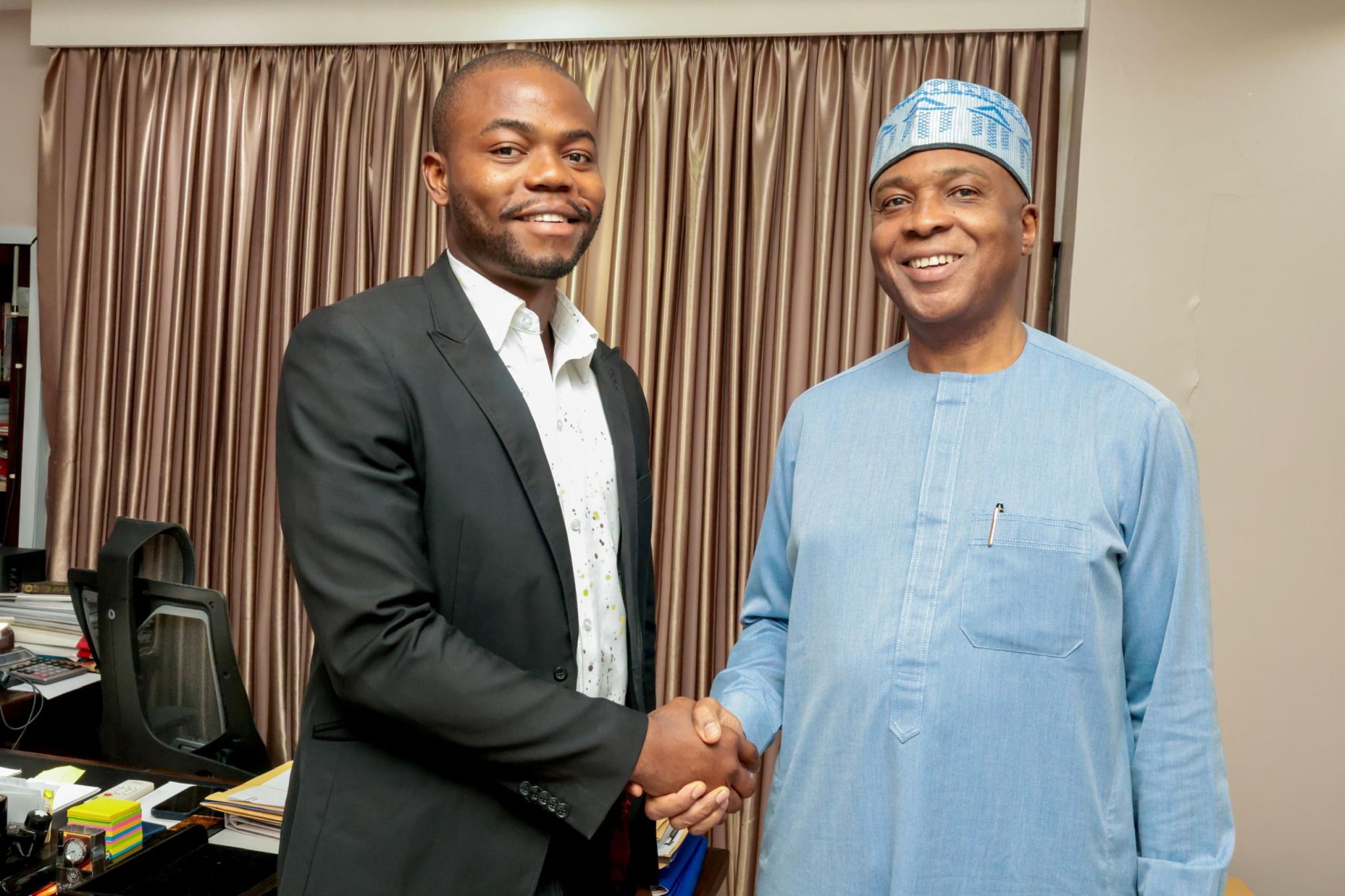 Israel Adeniyi Adekunle in a black suit shaking hands with Dr Abubakar Bukola Saraki, who is wearing a light blue traditional top and matching cap, in an office setting.
