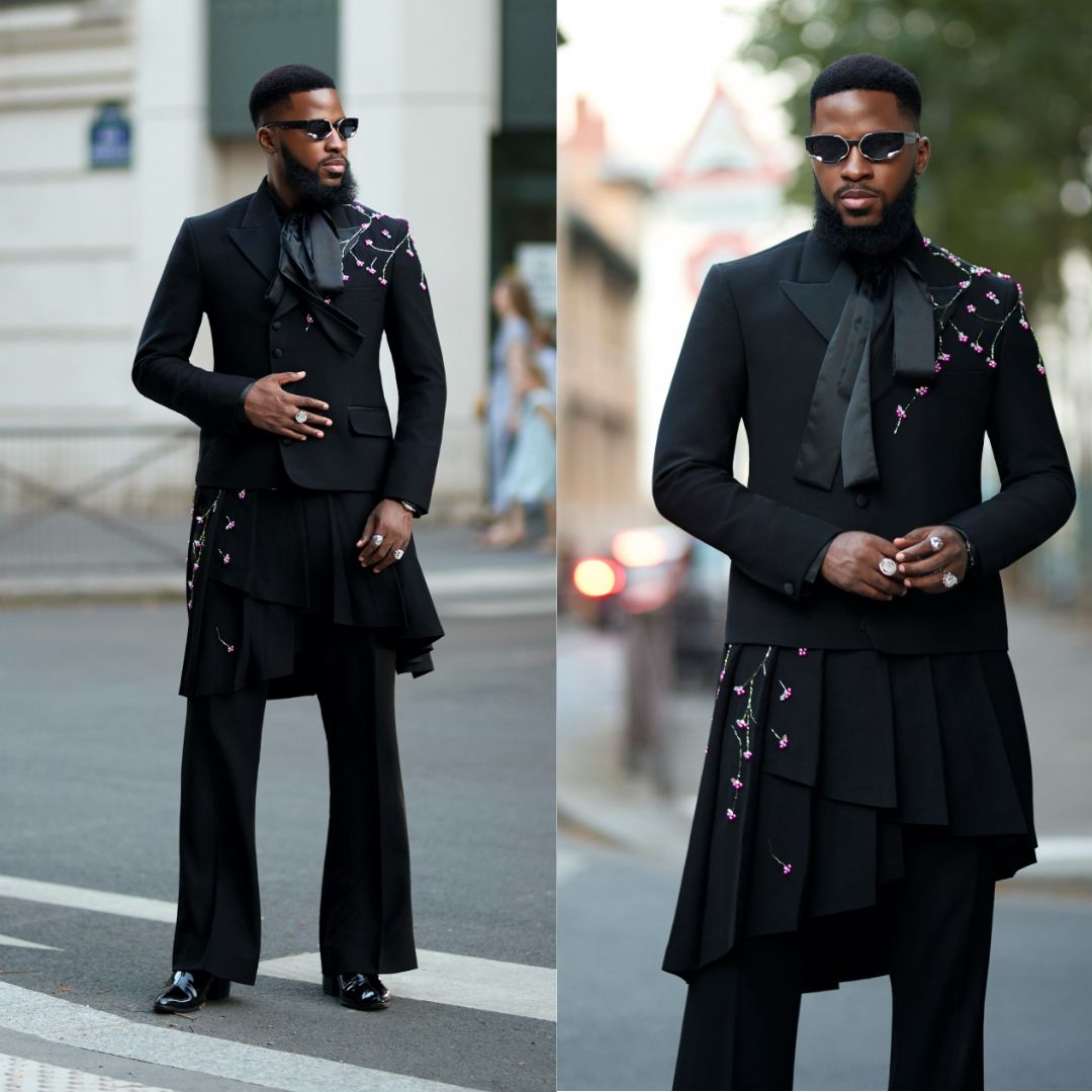 Akin Faminu standing on a street crosswalk in a black OJ Clothing cropped blazer with floral embroidery, an asymmetric tiered kilt, and wide-leg trousers.