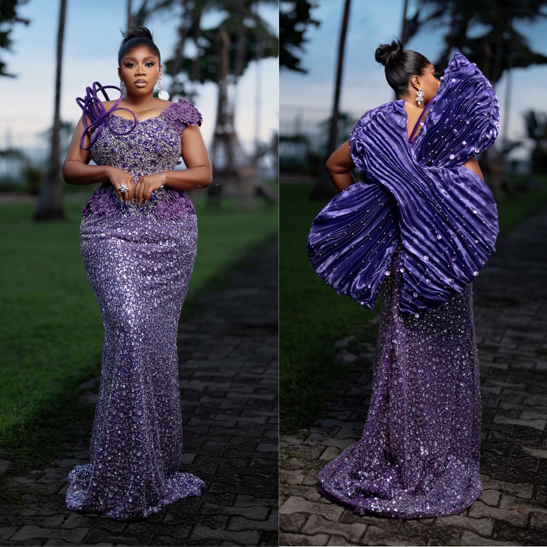 Wumi Toriola wearing a floor-length violet gown saturated with sequins and metallic beads, featuring an asymmetric neckline with floral applique on one shoulder and a structured, fan-like sculptural wing extending from the back.