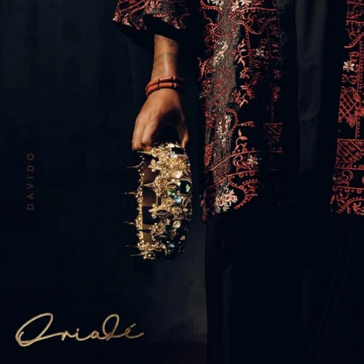 Official album cover art for Oriade by Davido featuring a close-up profile of an embroidered traditional garment and a hand holding a detailed gold crown adorned with green gemstones against a dark background.