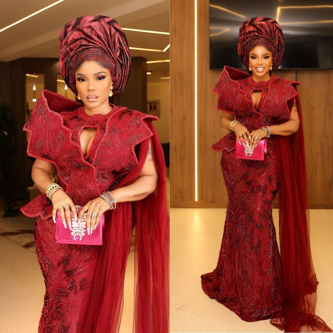 Iyabo Ojo wearing a deep burgundy beaded lace gown with structured architectural shoulder collar, keyhole neckline, trailing tulle cape, and pleated gele.