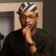 BBNaija alumnus Adekunle Olopade wearing a black traditional outfit, glasses, and a striped black and white cap.