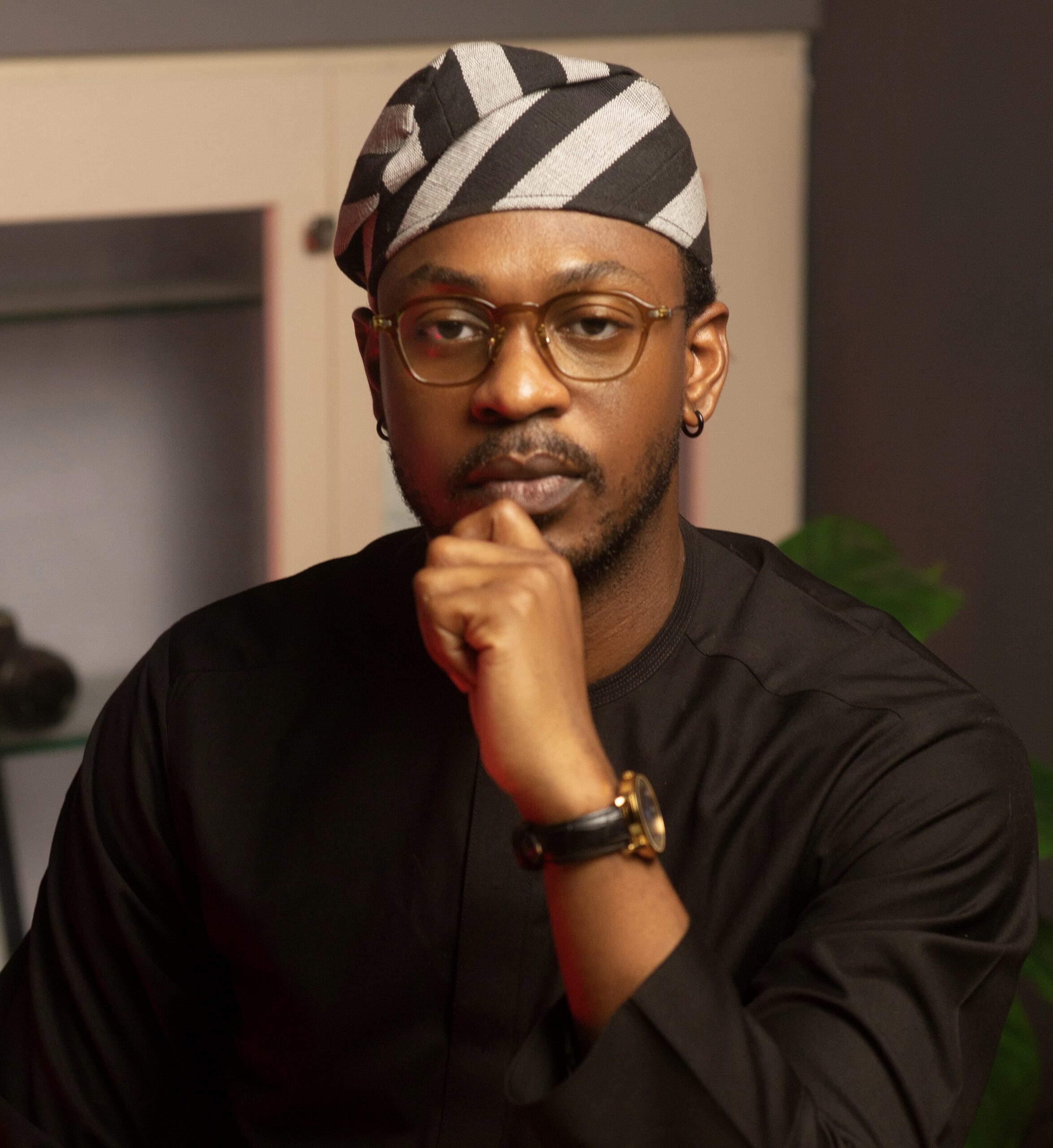 BBNaija alumnus Adekunle Olopade wearing a black traditional outfit, glasses, and a striped black and white cap.