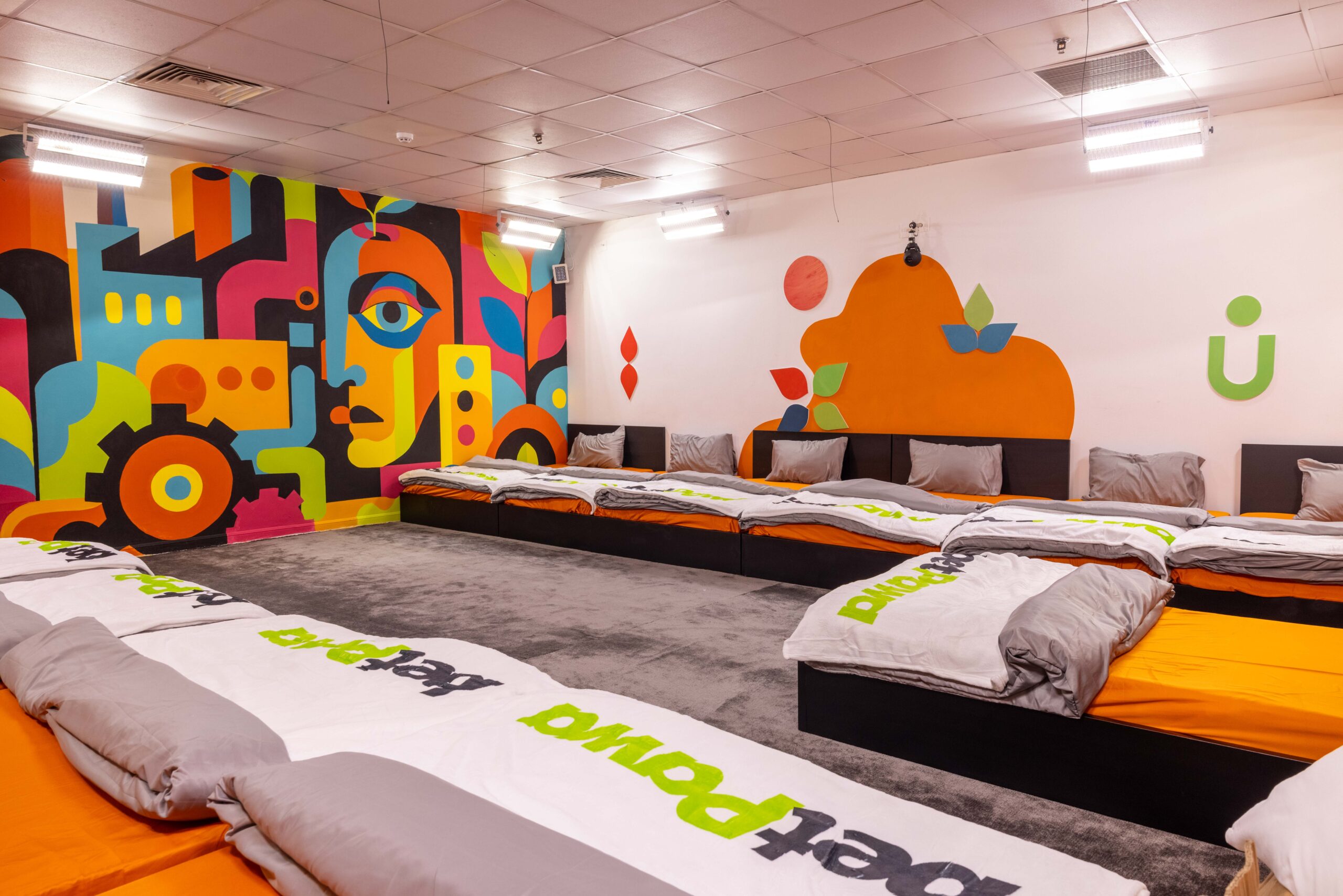 Housemates' bedroom in the Big Brother Naija Season 11 house featuring orange bedding, grey pillows, and a colourful geometric face mural