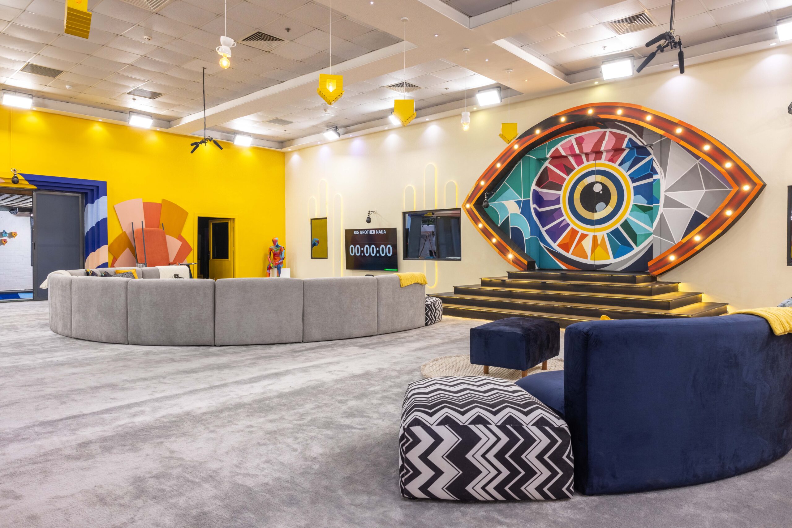 Main lounge in the Big Brother Naija Season 11 house featuring a large colourful illuminated Big Brother eye stage setup and digital countdown clock on