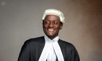 Nehemiah Shanum Danjuma in a black suit, white legal neckbands, and a traditional blonde-white barrister wig next to a hanging ceremonial gown.