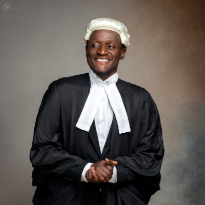 Nehemiah Shanum Danjuma in a black suit, white legal neckbands, and a traditional blonde-white barrister wig next to a hanging ceremonial gown.