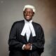 Nehemiah Shanum Danjuma in a black suit, white legal neckbands, and a traditional blonde-white barrister wig next to a hanging ceremonial gown.