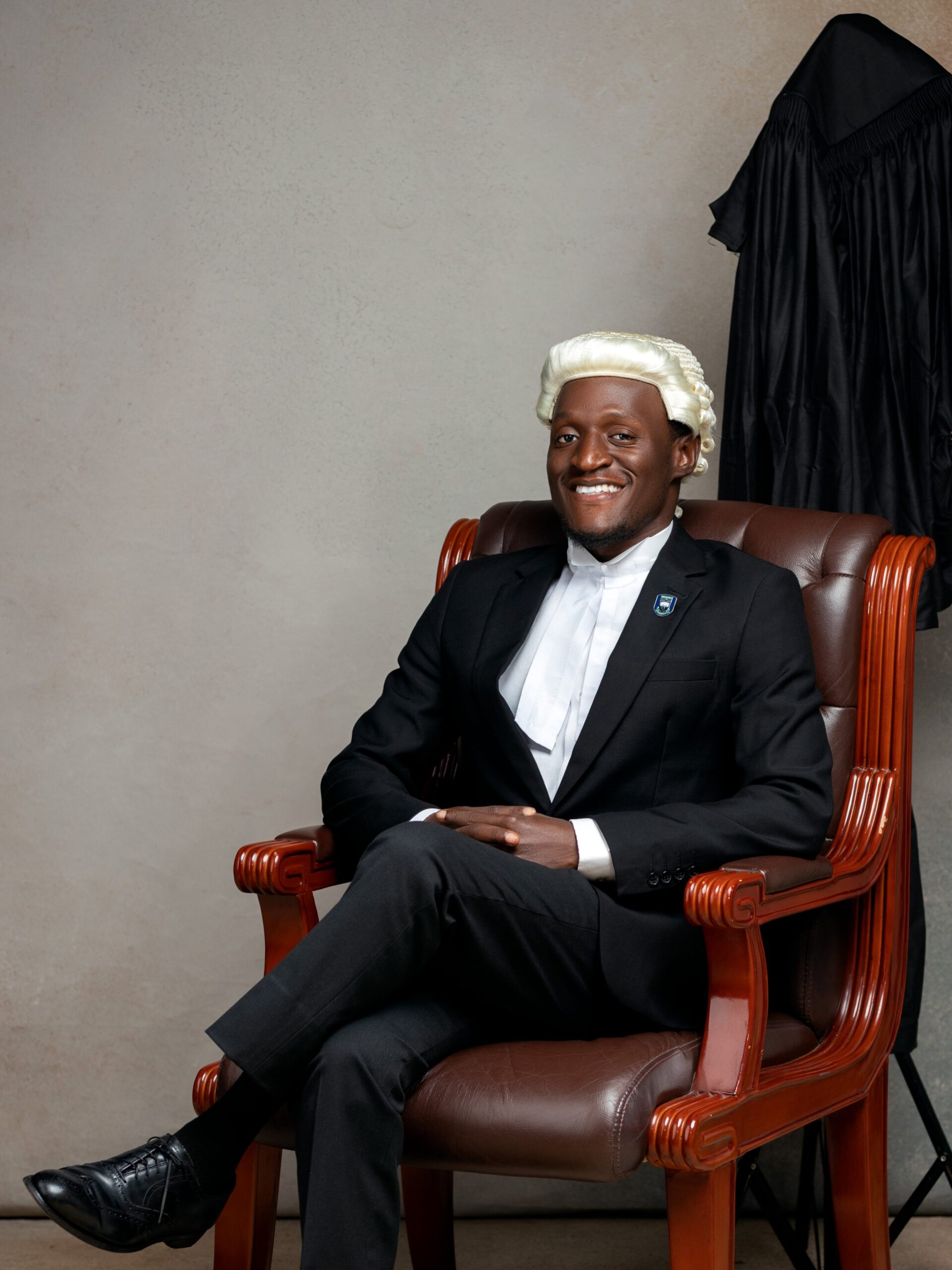 Nehemiah Shanum Danjuma, Esq. in a black corporate suit and legal wig, smiling while seated in a brown leather armchair.