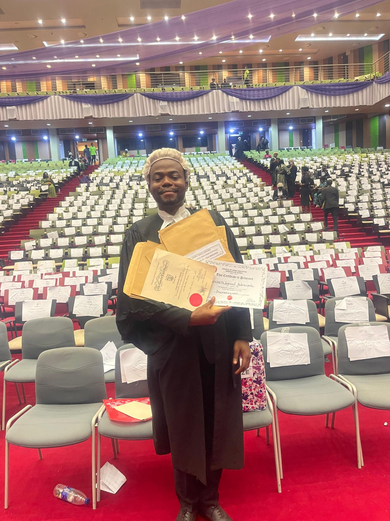 Adeniyi Adekunle standing in his barrister wig and gown inside the ceremony hall, holding his official Nigerian Law School certificate alongside multiple brown academic prize envelopes.