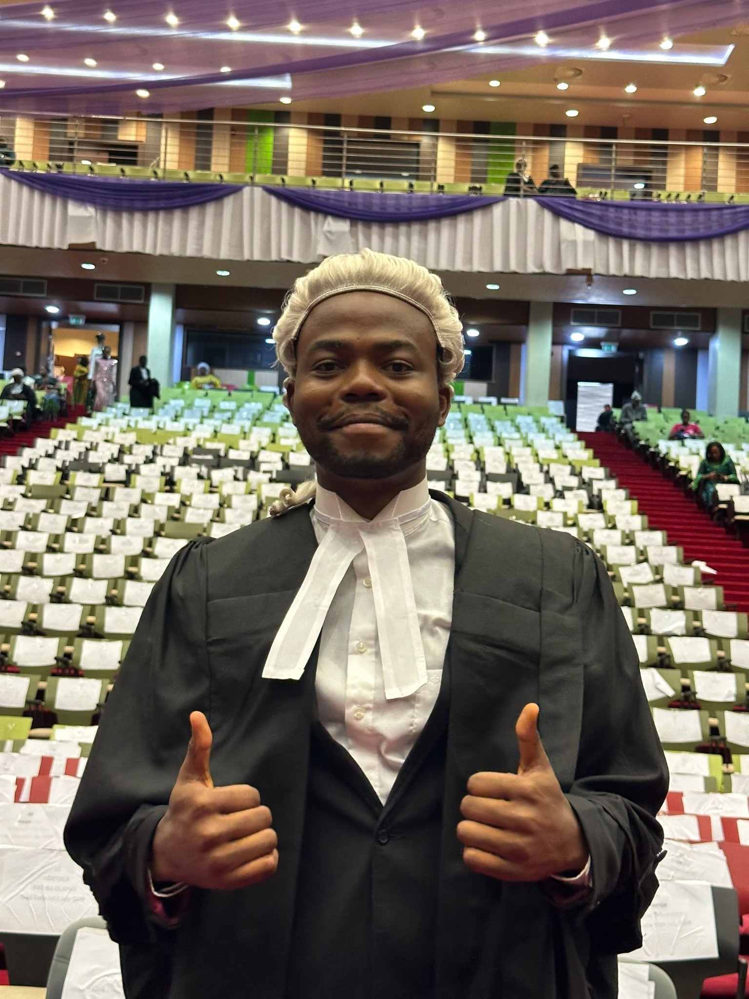 Adeniyi Adekunle smiling and giving a double thumbs-up while wearing his legal practitioner wig, black gown, and white collar bands in the Call to Bar auditorium.