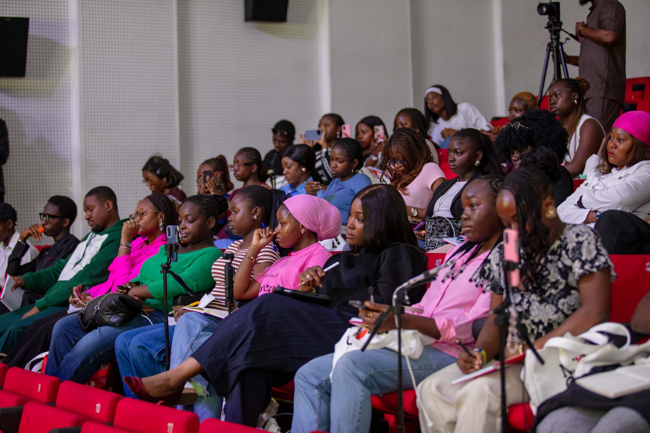 A side angle view of a row of young African content creators sitting in red auditorium seats, attentively listening and taking notes during a presentation.