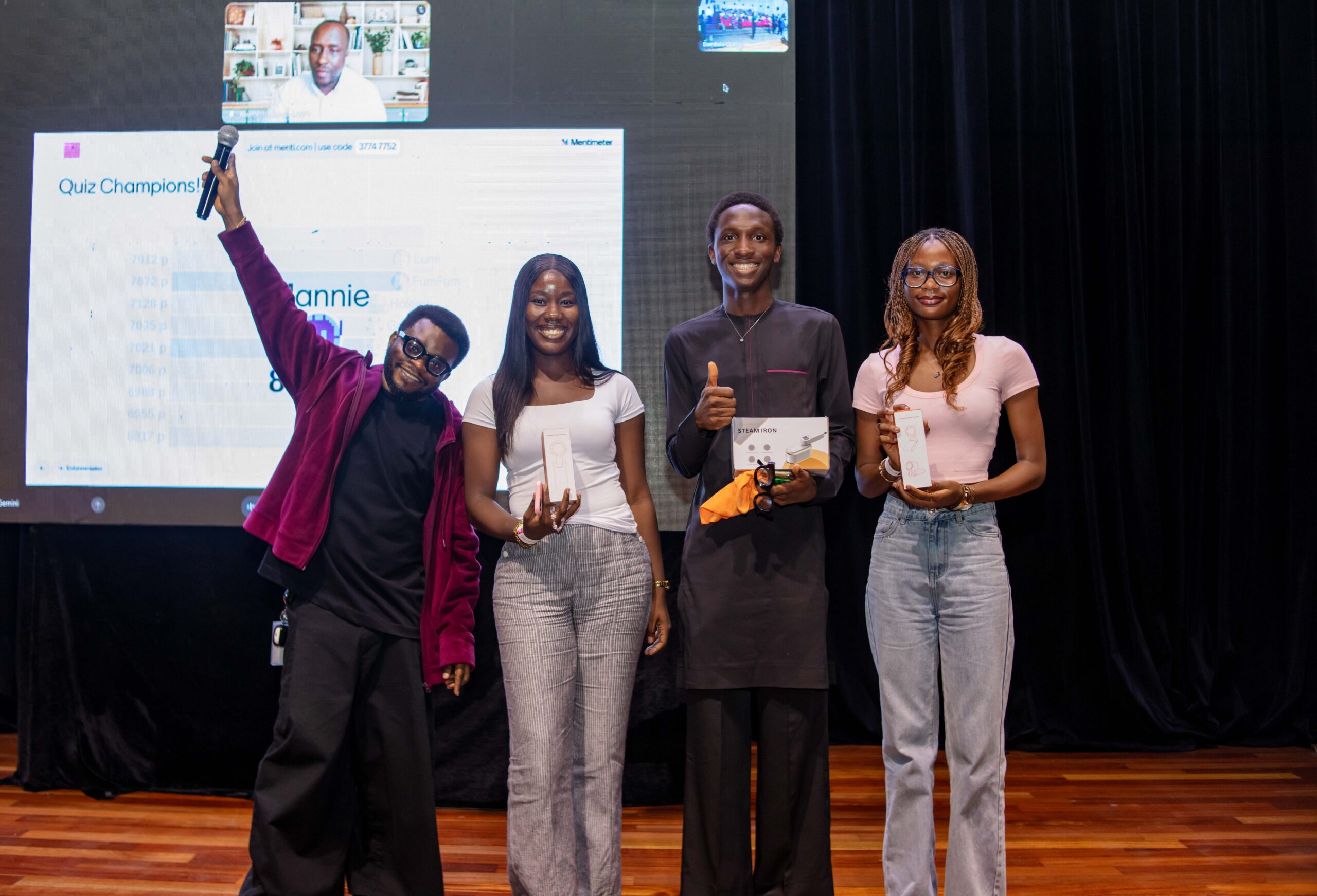 Taiwo Kola-Ogunlade celebrating on stage next to three young creative challenge winners holding boxed rewards and digital prizes.