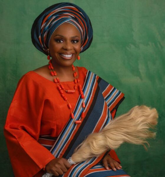 Nigerian author Lola Shoneyin smiling in an orange iro and buba with a striped multi-coloured aso-oke gele, shoulder sash, and a traditional irukere horsetail whip.