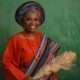 Nigerian author Lola Shoneyin smiling in an orange iro and buba with a striped multi-coloured aso-oke gele, shoulder sash, and a traditional irukere horsetail whip.
