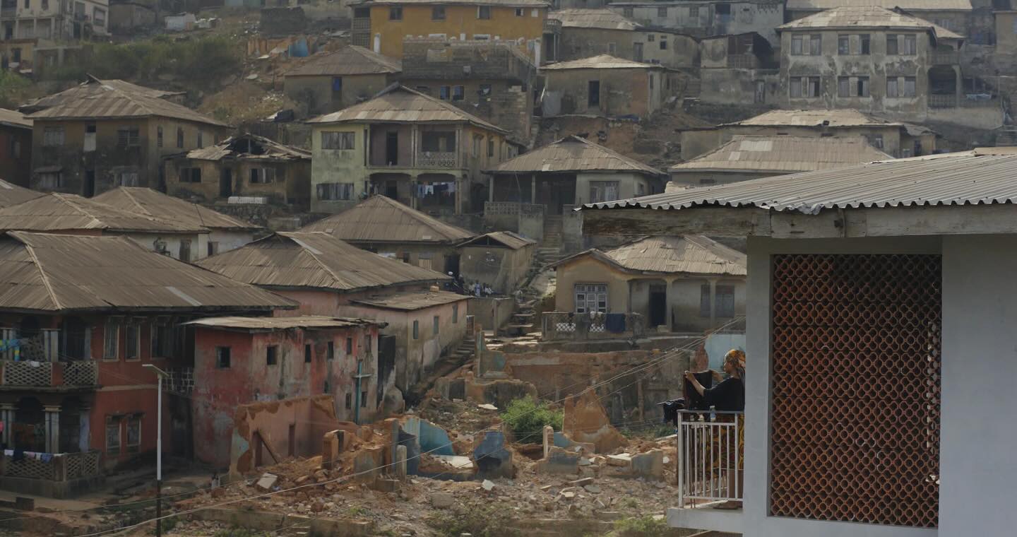 Teniola Aladese standing on a residential balcony looking out at old houses built on a hill in Ibadan Nigeria.