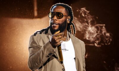 Burna Boy wearing tinted sunglasses, a beige blazer, a white t-shirt, and textured trousers holds a golden microphone under dramatic stage lighting with smoke elements.