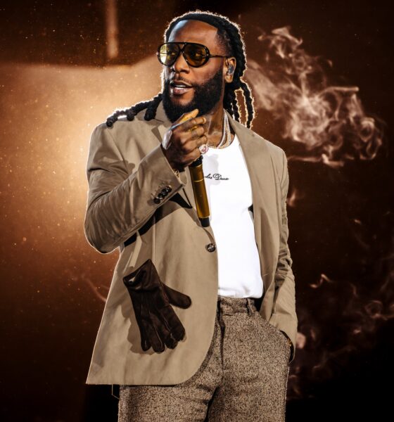 Burna Boy wearing tinted sunglasses, a beige blazer, a white t-shirt, and textured trousers holds a golden microphone under dramatic stage lighting with smoke elements.