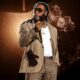 Burna Boy wearing tinted sunglasses, a beige blazer, a white t-shirt, and textured trousers holds a golden microphone under dramatic stage lighting with smoke elements.