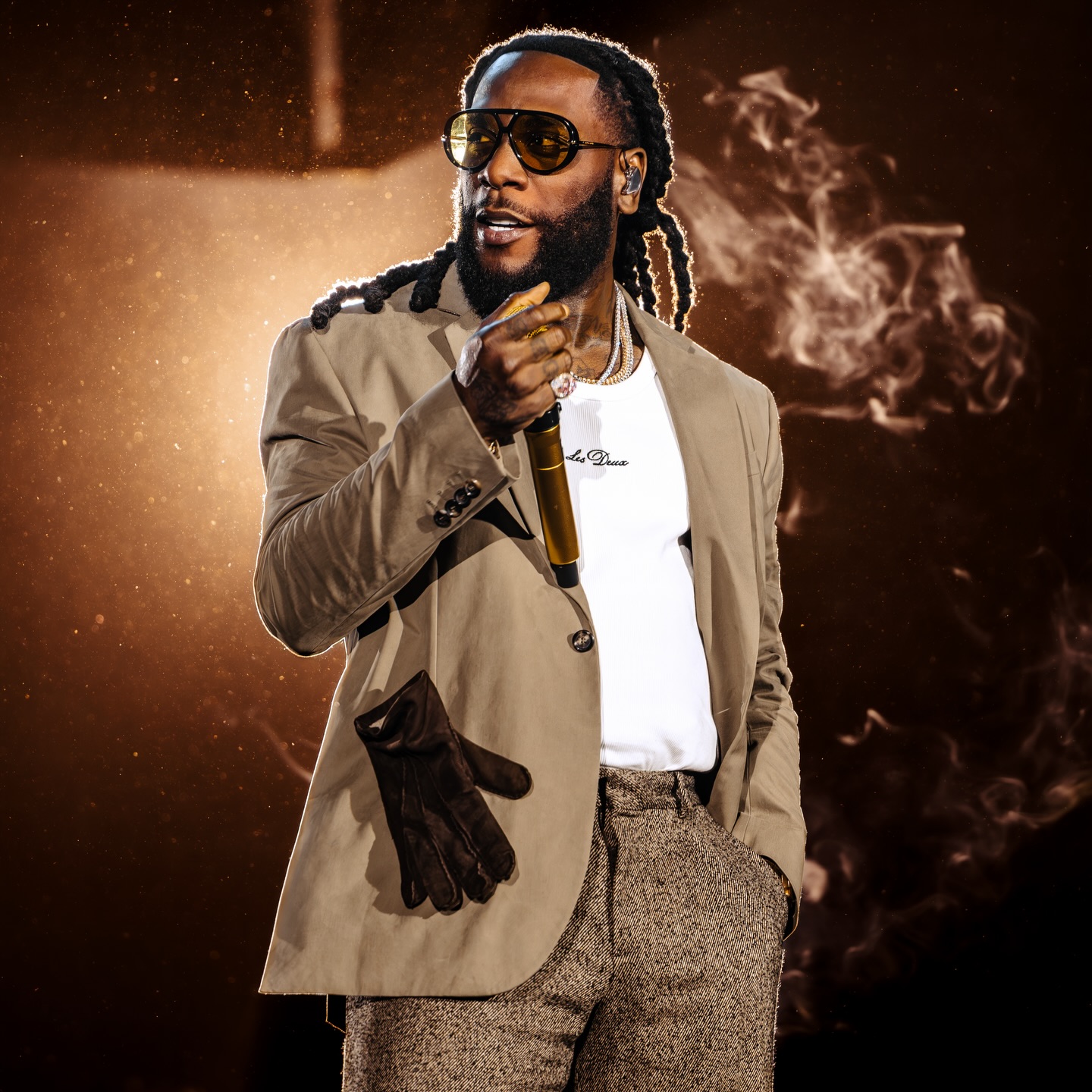 Burna Boy wearing tinted sunglasses, a beige blazer, a white t-shirt, and textured trousers holds a golden microphone under dramatic stage lighting with smoke elements.