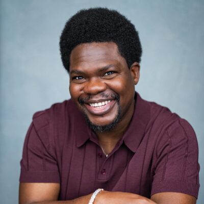 Nigerian actor Debo Adedayo, professionally known as Mr Macaroni, smiling in a maroon polo shirt for his official acting headshot.