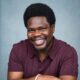Nigerian actor Debo Adedayo, professionally known as Mr Macaroni, smiling in a maroon polo shirt for his official acting headshot.