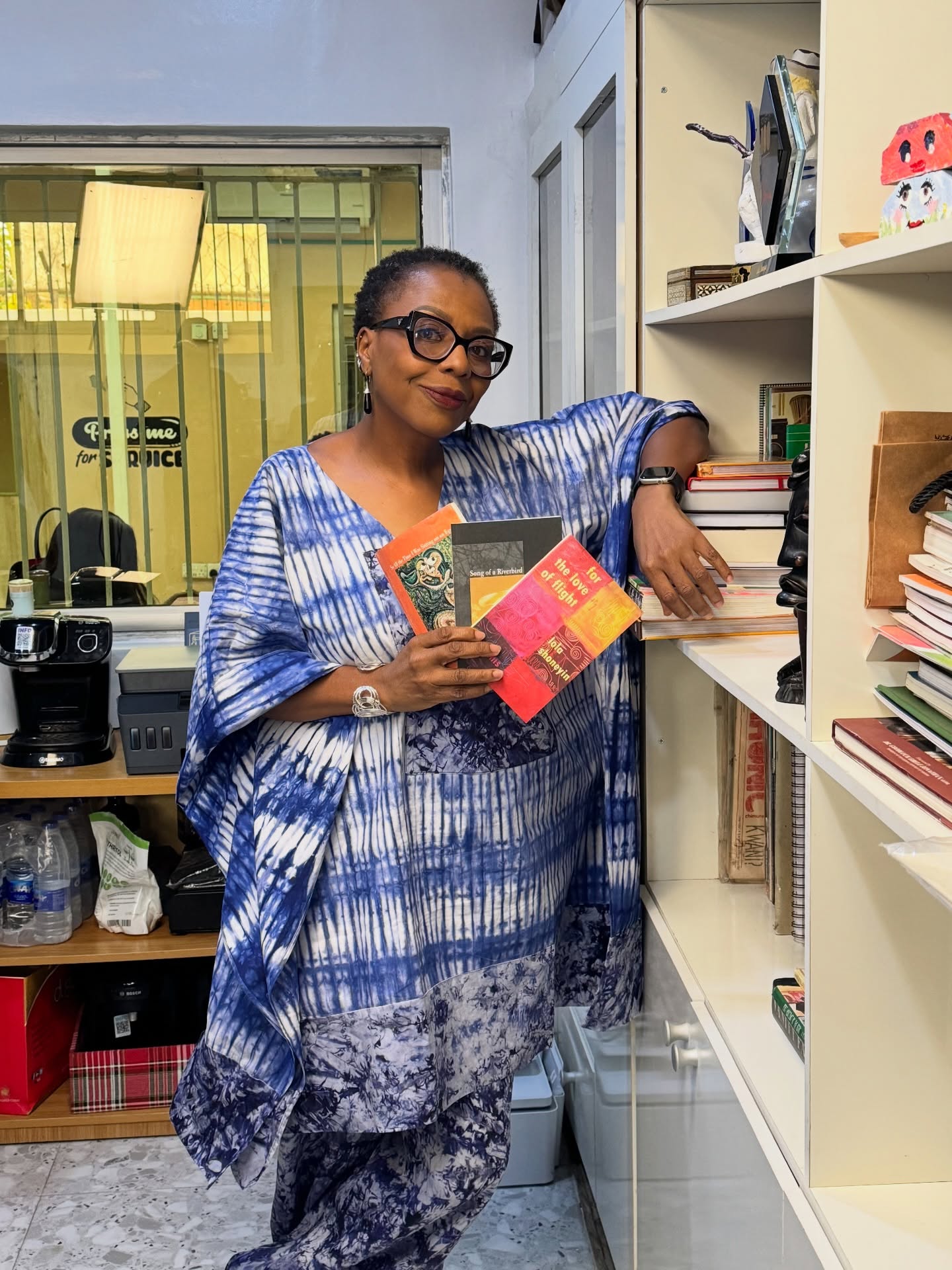 Nigerian author Lola Shoneyin wearing a blue adire kaftan while holding copies of her poetry books beside an office bookcase.