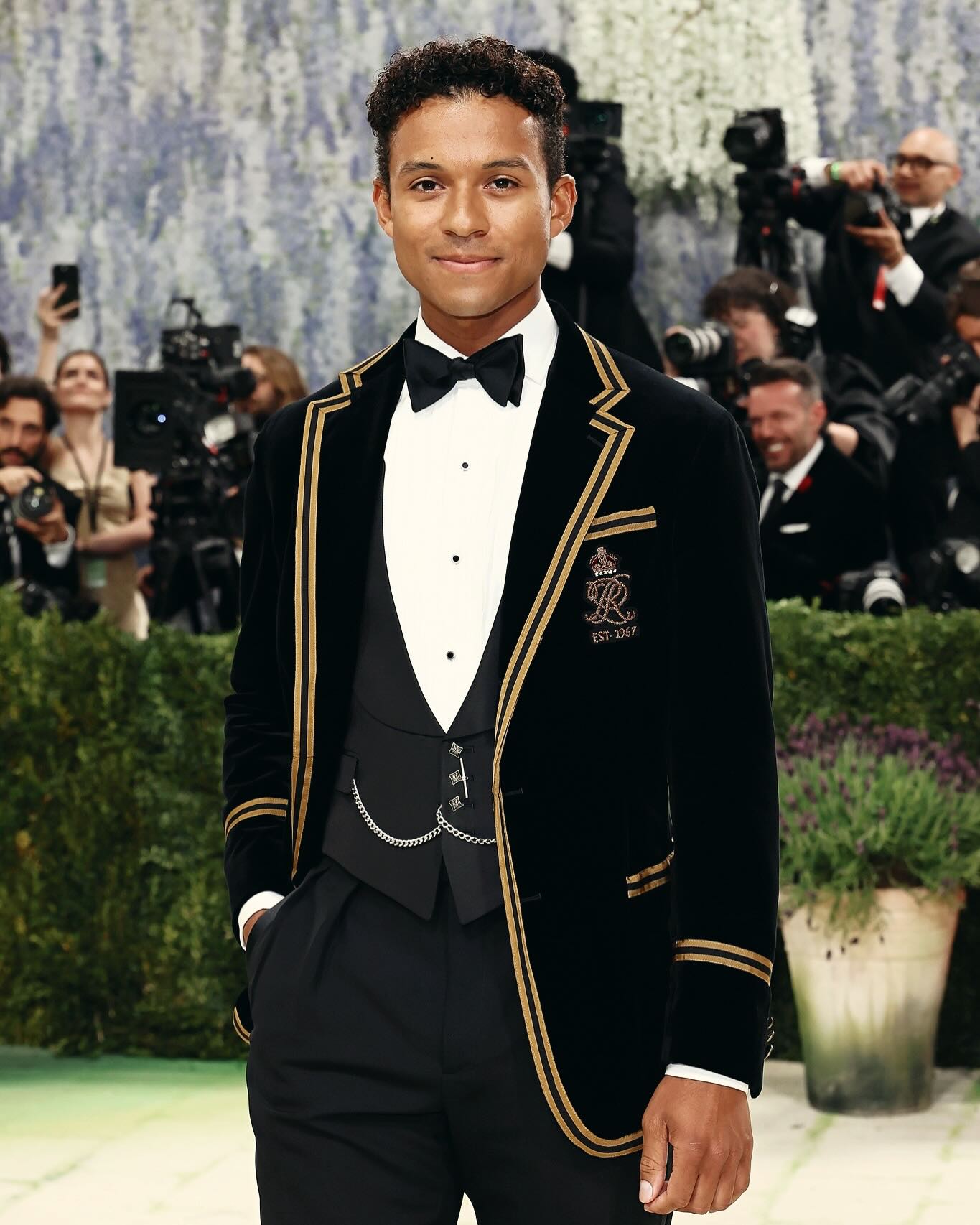 Actor Jaafar Jackson posing at the Met Gala wearing a black velvet Ralph Lauren cricket blazer with gold-taped trim, an embroidered crest, a white pleated tuxedo shirt, a black bow tie, and a pocket watch with a hanging silver chain