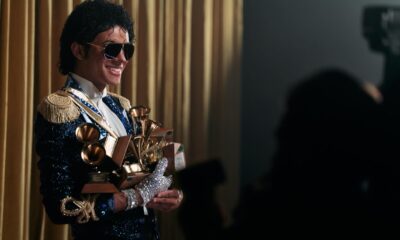 Jaafar Jackson as Michael Jackson wearing aviator sunglasses and a blue sequined military jacket with gold epaulettes, holding eight gold Grammy Awards.