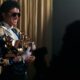 Jaafar Jackson as Michael Jackson wearing aviator sunglasses and a blue sequined military jacket with gold epaulettes, holding eight gold Grammy Awards.