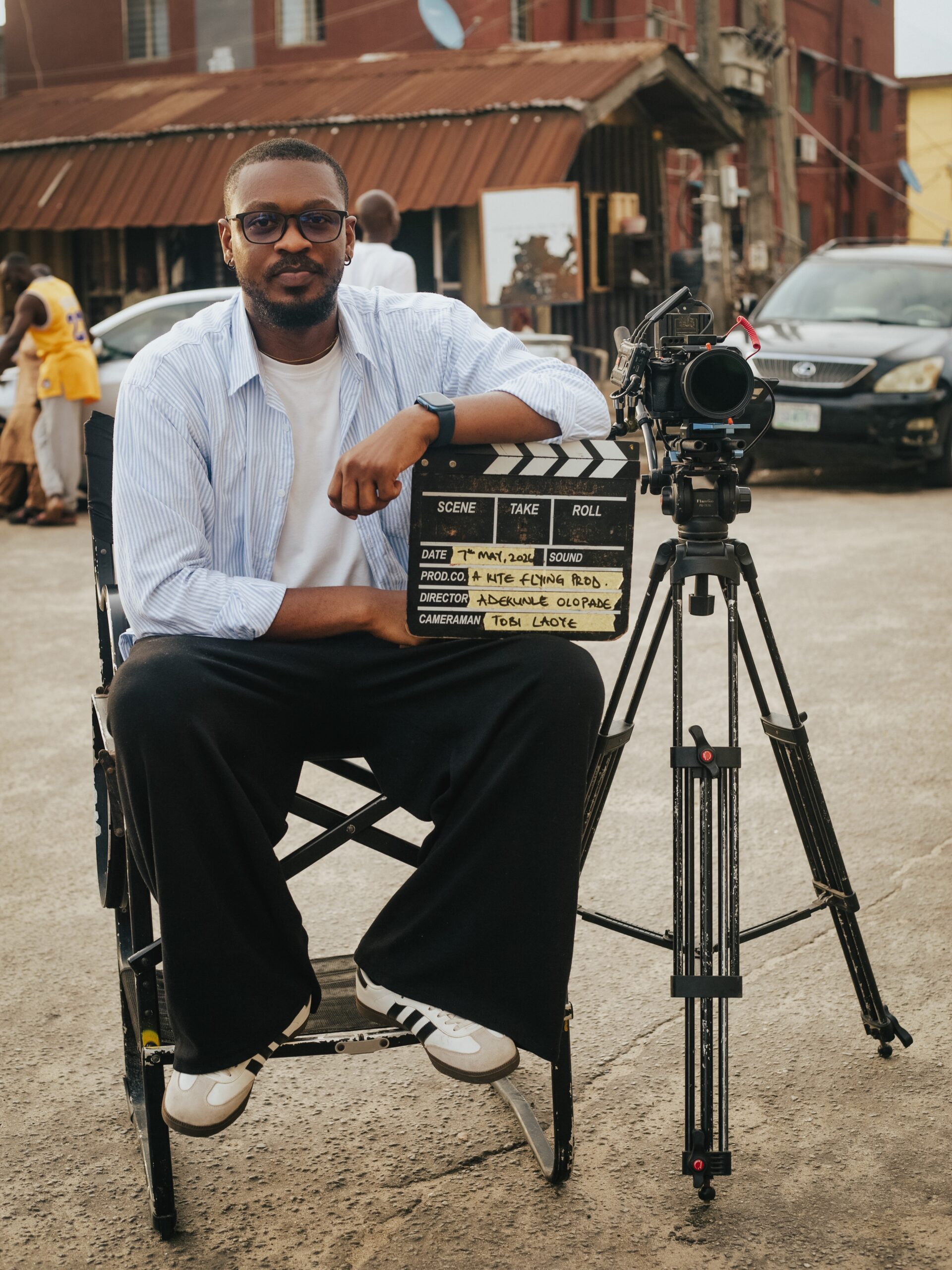 Adekunle Olopade sitting in a director chair on an outdoor movie set holding a movie clapperboard next to a video camera on a tripod.