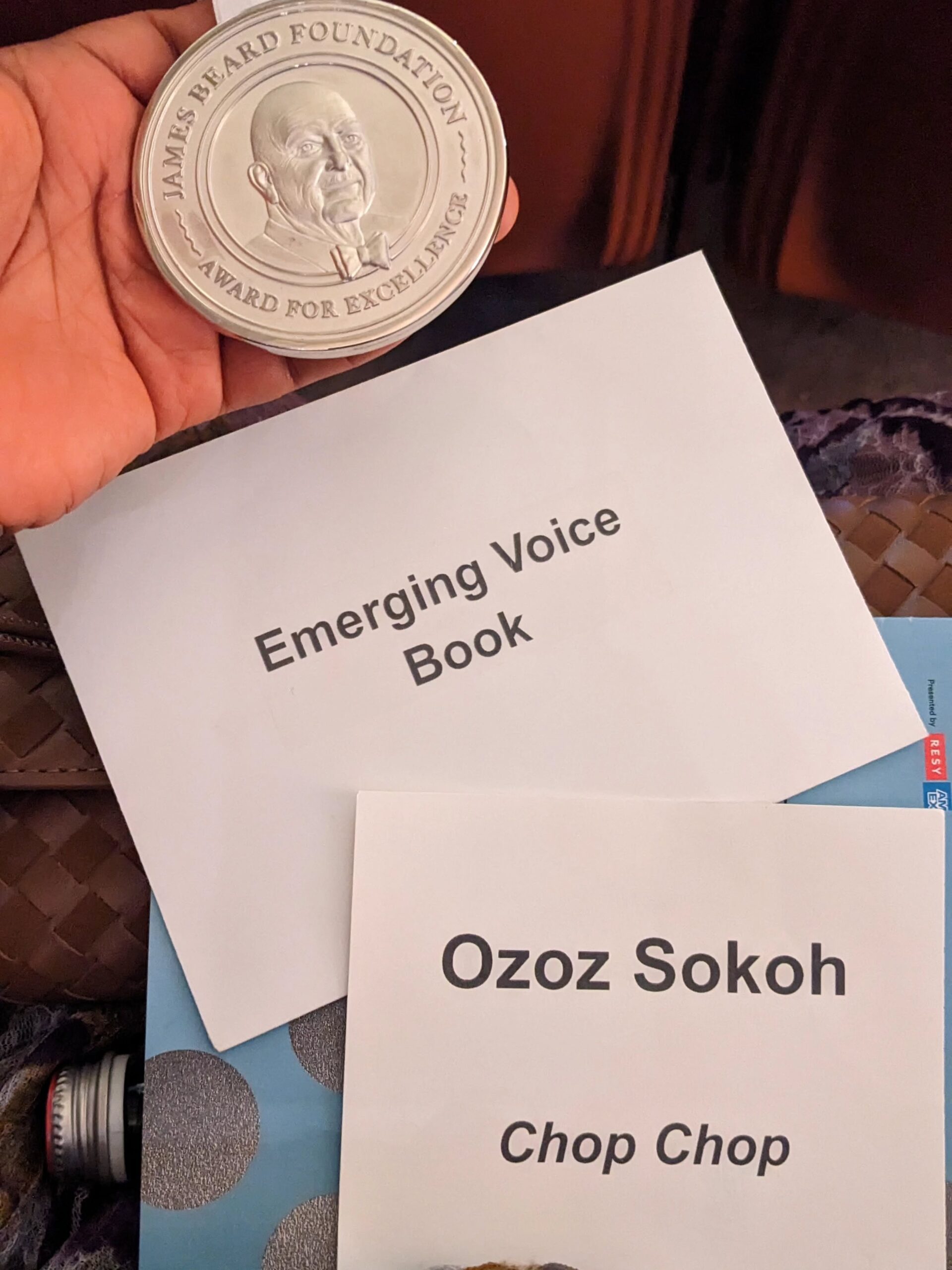 A silver James Beard Foundation Award for Excellence medal held over custom name cards reading Ozoz Sokoh, Chop Chop, and Emerging Voice Book.