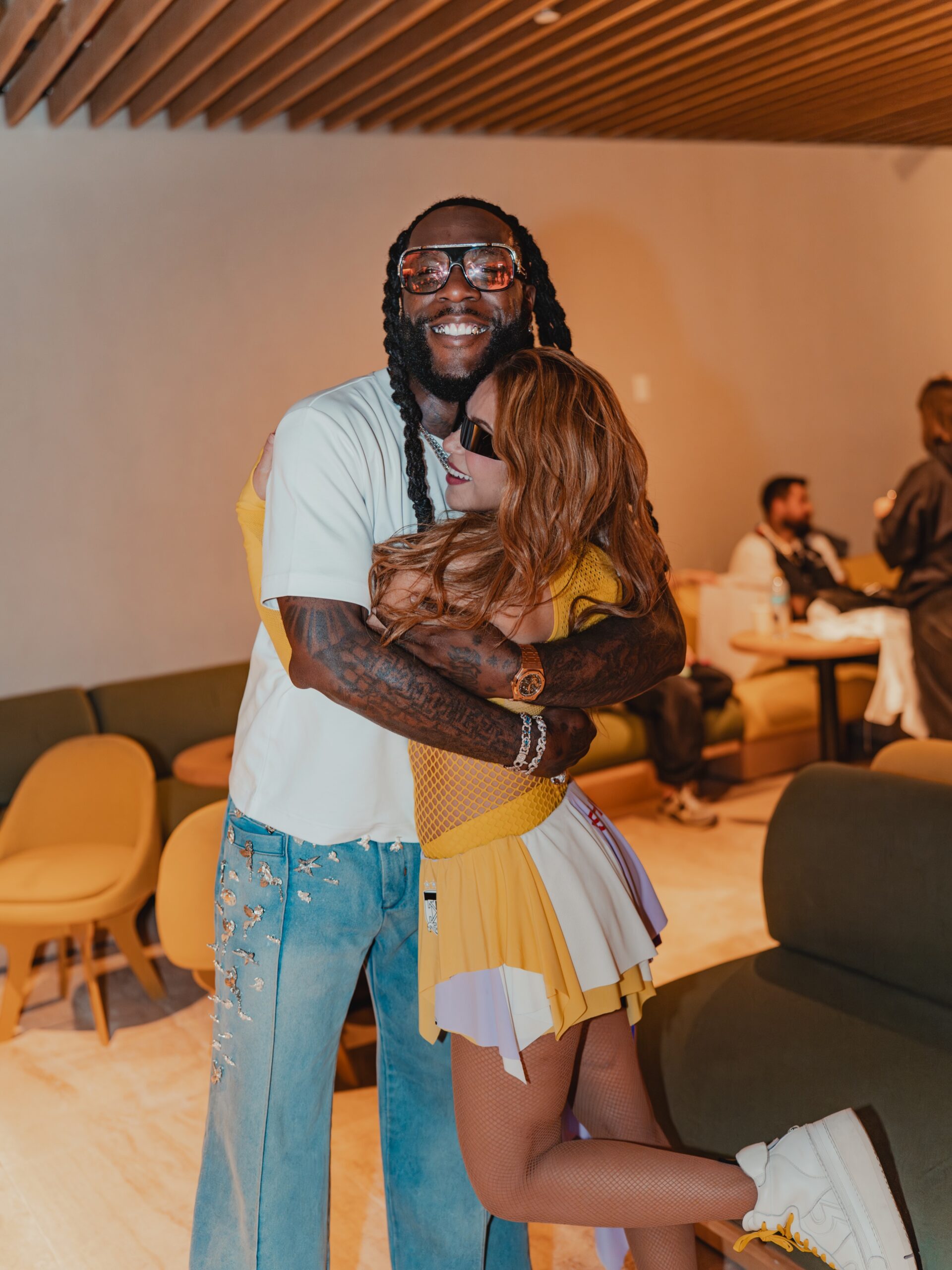 Nigerian artist Burna Boy in a white t-shirt and jeans smiles whilst hugging Colombian singer Shakira, who wears a yellow and white performance outfit and sunglasses.