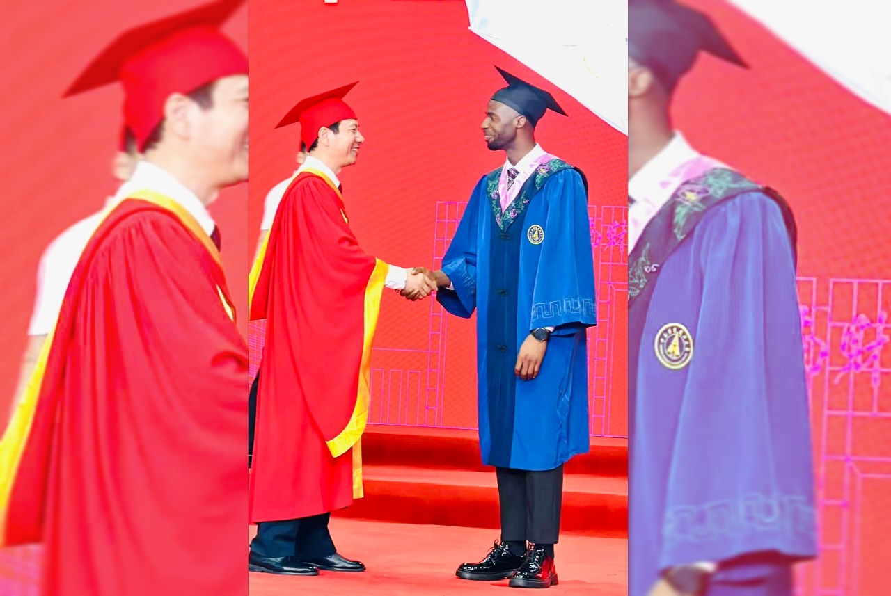 Nigerian scholar Nnabuike Chisom in blue academic regalia shaking hands with a university official in a red robe on stage.