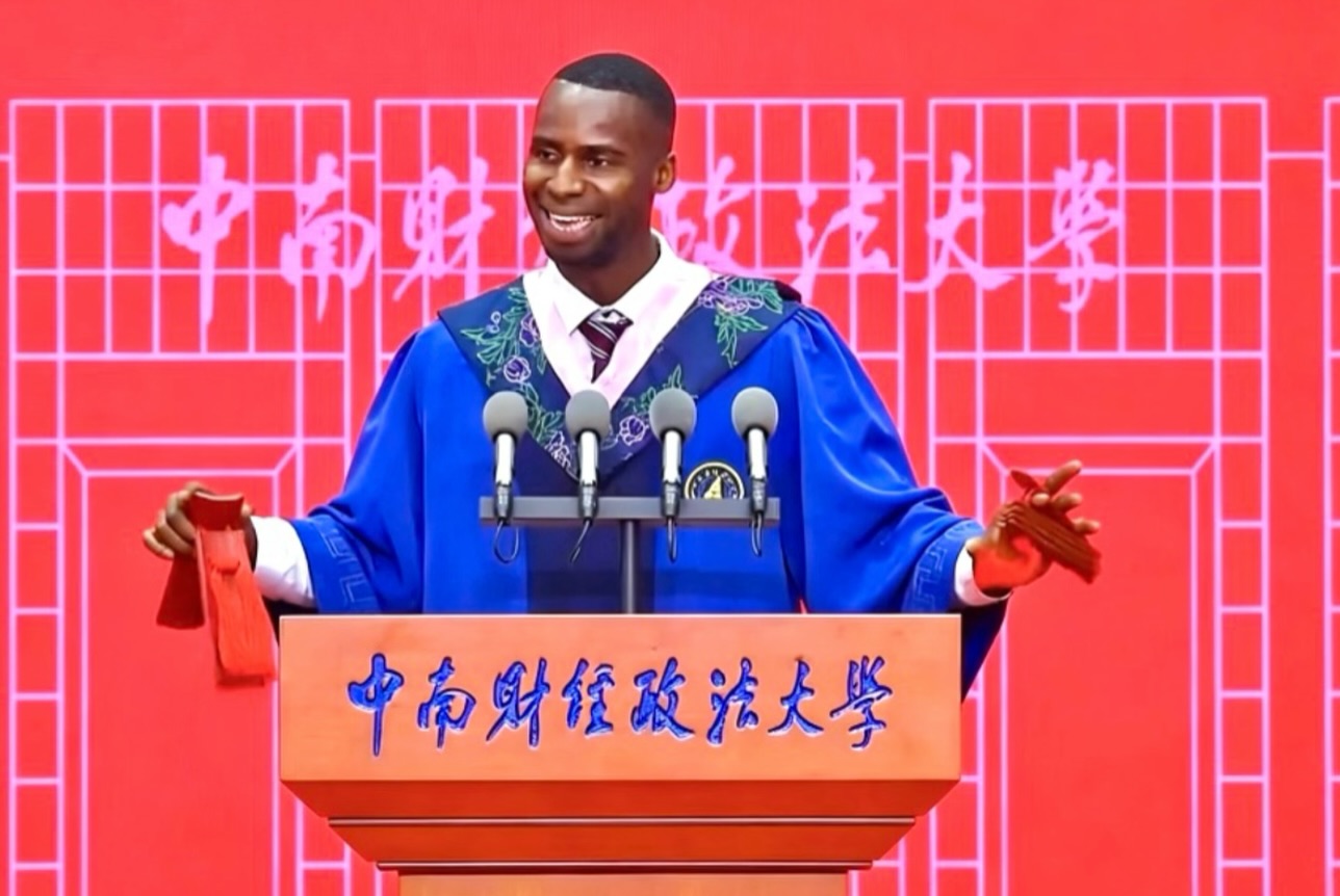 Nnabuike Chisom smiling at the podium while wearing blue and navy embroidered Chinese graduation robes and holding a red tassel.