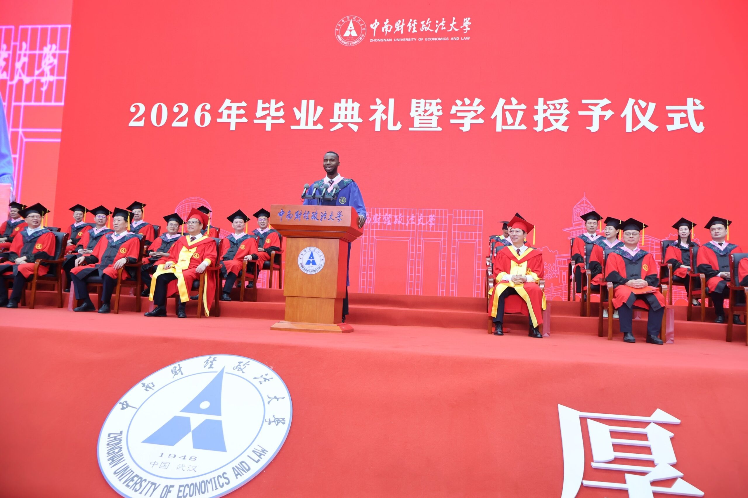 Nnabuike Chisom delivers his keynote address at a wooden podium on stage before a panel of university faculty members.