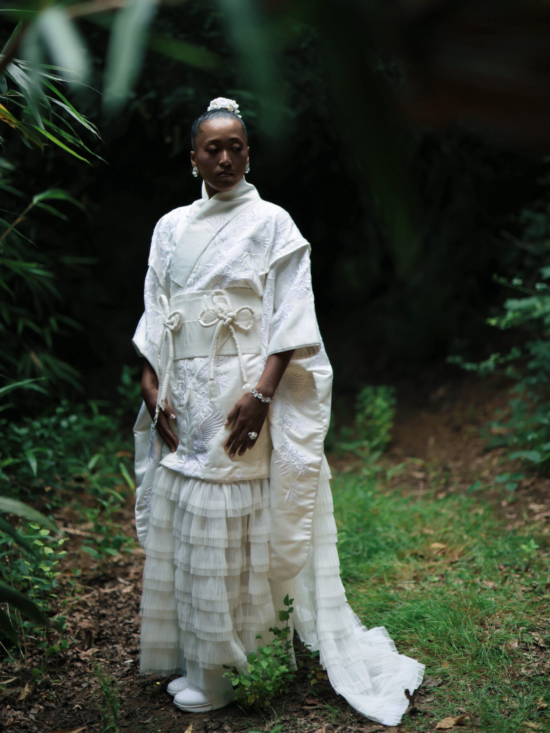 Full-length portrait of Naomi Osaka in a forest wearing a structured white obi belt and embroidered robe from Hana Yagi's Evolving Ceremony collection.