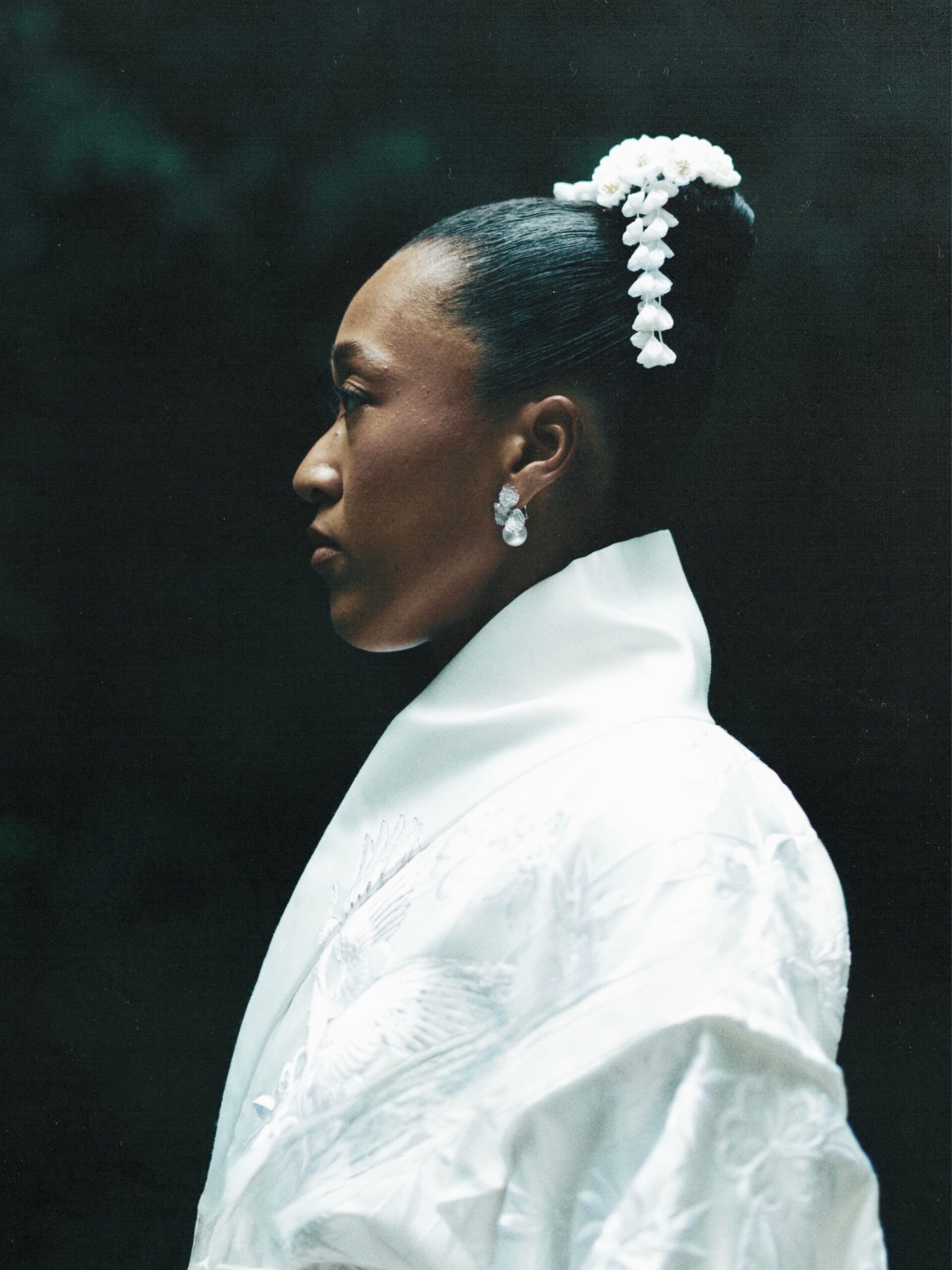 Side profile portrait of Naomi Osaka wearing a high-collared white ceremonial dress against a dark backdrop.