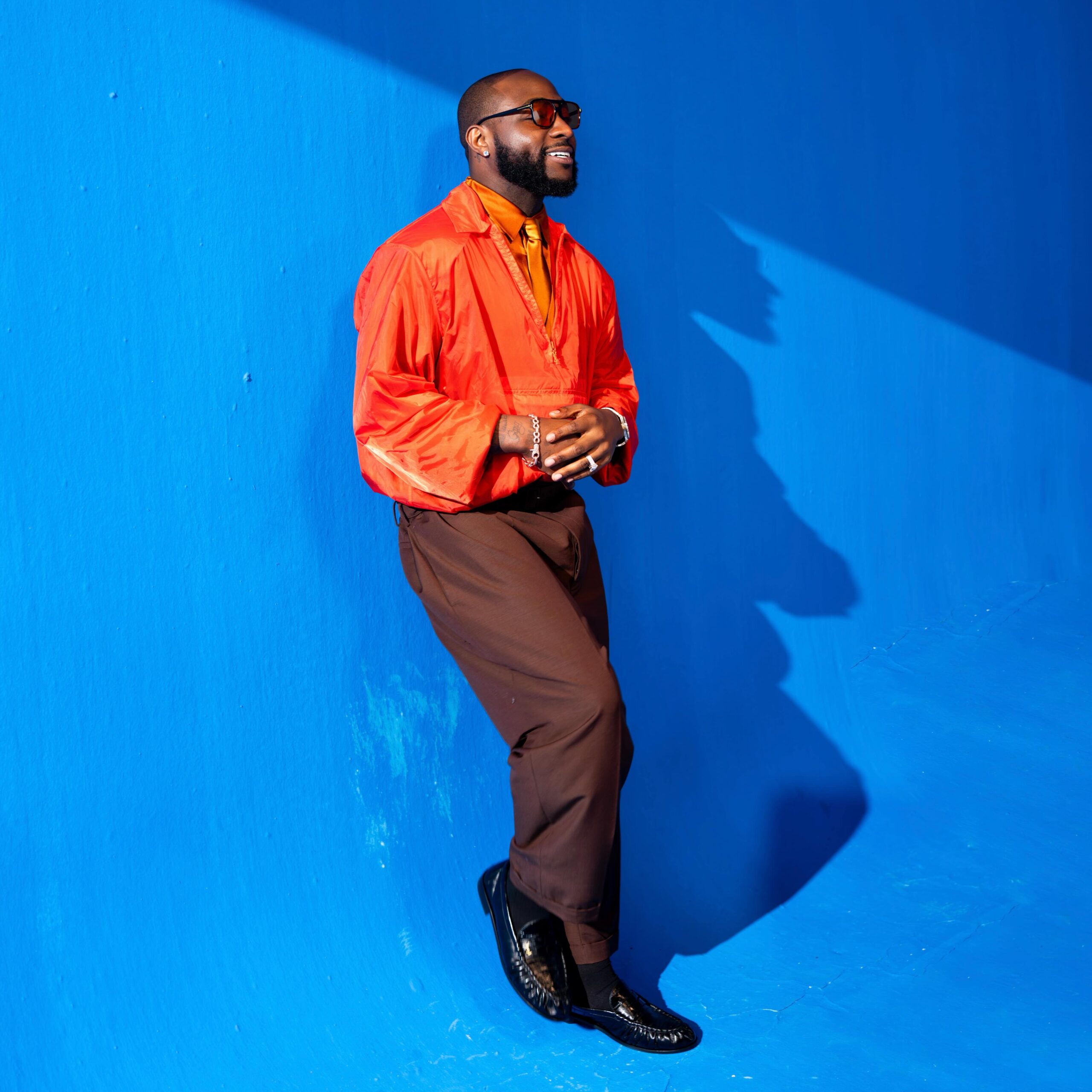 Music video still of Davido smiling while leaning against a vibrant blue studio wall, wearing a bright orange zip-up pullover jacket, brown trousers, sunglasses, and a gold tie from the I Know Who I Be visuals.